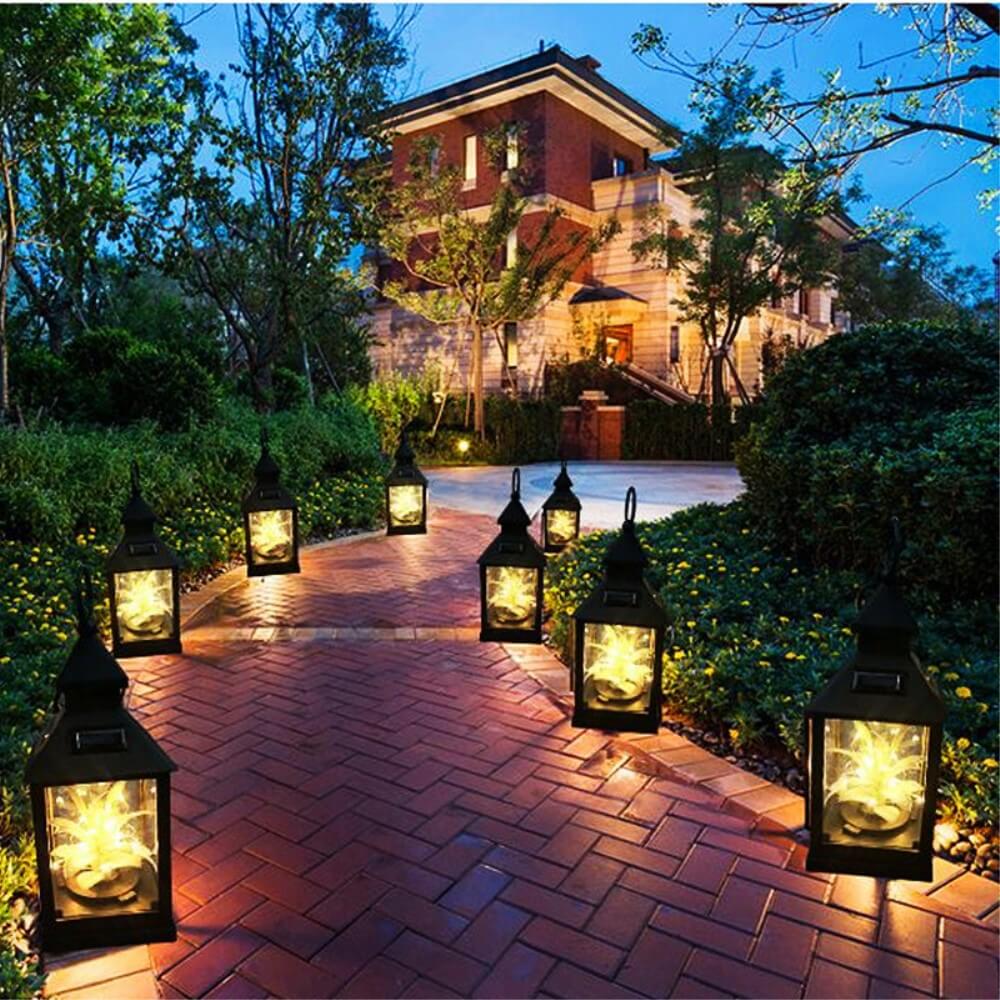 Ein gepflasterter Weg, gesäumt von leuchtenden Cover-Discount Solar Tischlampe LED Laternen, die warmweißes Licht verbreiten, führt zu einem großen Haus, das in der Abenddämmerung von Bäumen umgeben ist.