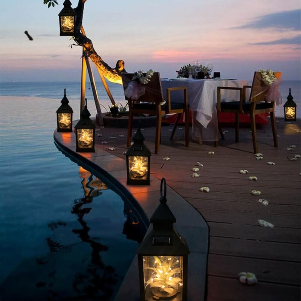 Ein romantisches Abendessen im Freien am Pool bei Sonnenuntergang, beleuchtet vom warmweißen Licht der Cover-Discount Solar Tischlampe LED Laterne, mit auf dem Deck verstreuten Laternen und Blütenblättern.