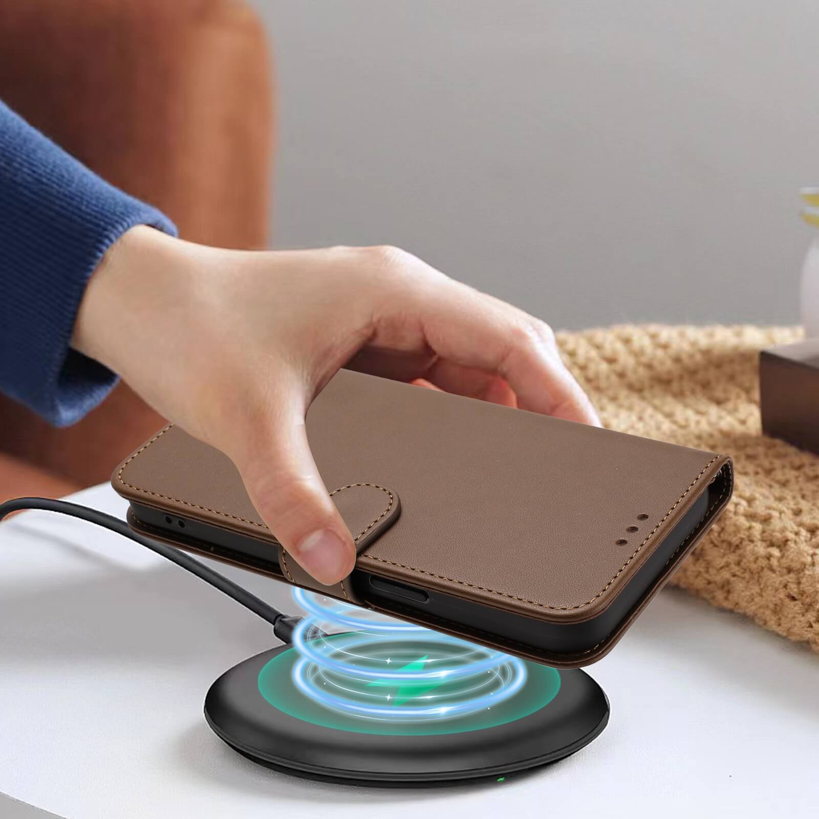 Eine Hand legt ein Cover-Discount iPhone 17 Pro Glänzendes Hüllen Etui mit Handschlaufe in braunem PU-Leder auf ein kabelloses Ladepad auf dem Tisch.
