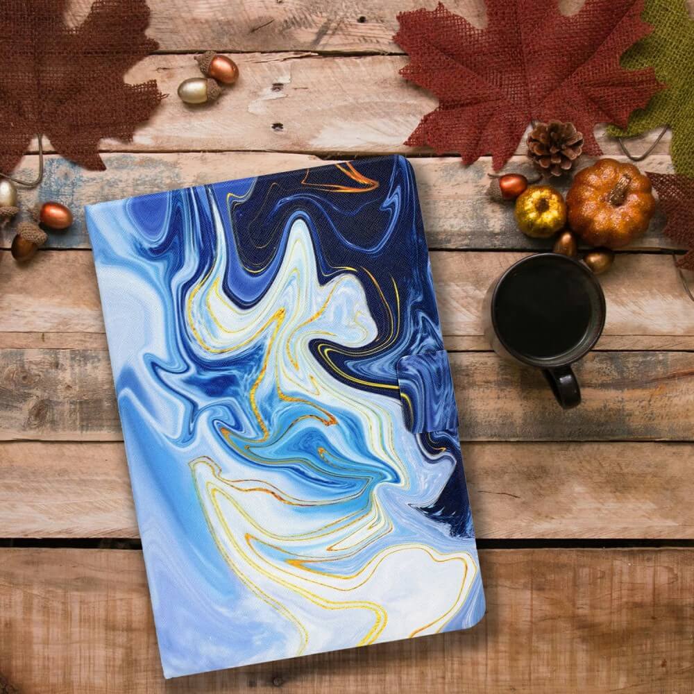 Ein blau-weiß marmoriertes Notizbuch liegt neben Kaffee, Kürbissen, Blättern, Eicheln und einem Cover-Discount Kindle Paperwhite 2024 Etui mit stilvollem Motiv auf einem Holztisch.