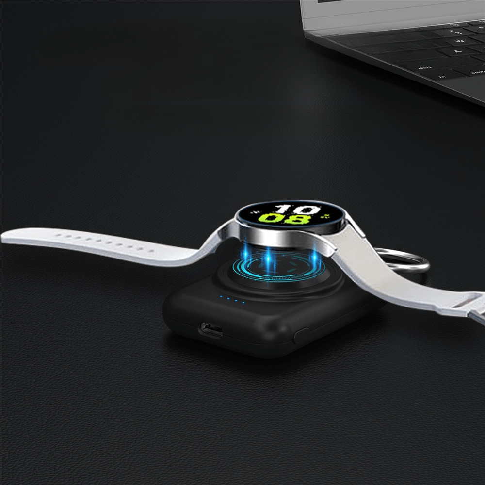 Eine Cover-Discount.ch Mini-Powerbank Samsung Galaxy Watch lädt eine Smartwatch über kabelloses Laden auf einem schwarzen Pad, neben einem Laptop auf einer dunklen Oberfläche - und bietet 1800mAh mühelosen Komfort.