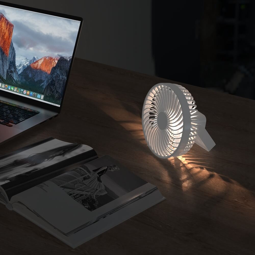 Ein kompakter weißer Mini-Ventilator von Cover-Discount für Camping steht neben einem offenen Laptop und einer Zeitschrift auf einem Holztisch und ist bei schwachem Licht sanft beleuchtet.