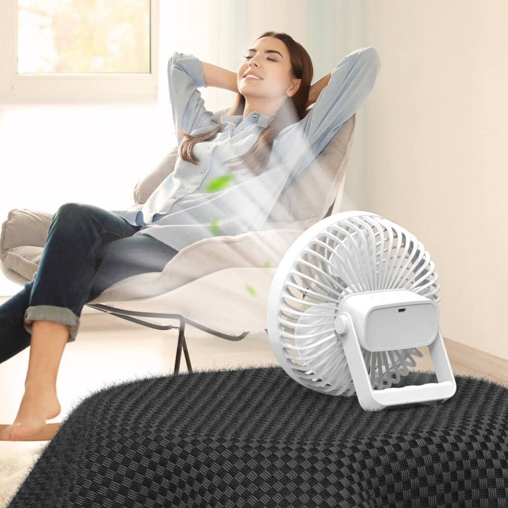 Eine Frau entspannt sich in einem Stuhl, während der tragbare Mini-Ventilator Camping von Cover-Discount ihr in einem hellen, gemütlichen Raum sanft kühle Luft zubläst.