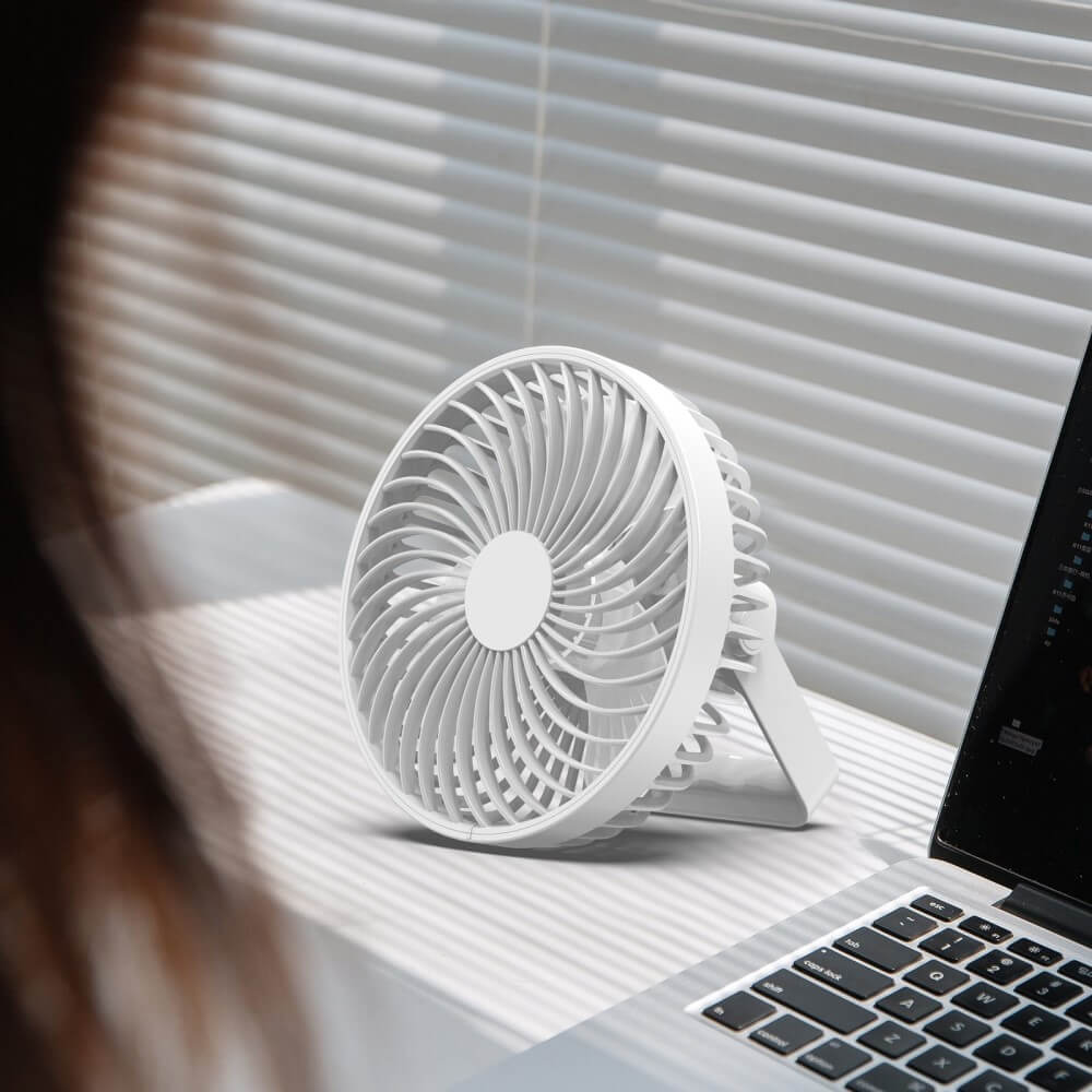 Ein weisser Mini-Ventilator von Cover-Discount für Camping steht auf einem Schreibtisch neben einem Laptop, im Hintergrund sind Jalousien zu sehen. Dieser tragbare Ventilator eignet sich ideal für die stilvolle und bequeme Kühlung des Arbeitsplatzes.