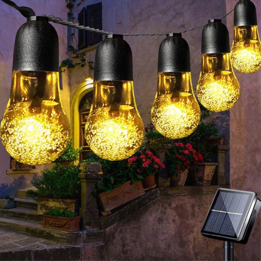 Die Cover-Discount Solar LED Lichterkette mit Glühbirnen wird im Außenbereich an Blumentöpfen und einer Treppe aufgehängt und sorgt für eine charmante Gartenbeleuchtung. Ein Solarpanel versorgt die warmen LED-Lichter mit Strom für gemütliche Abende.
