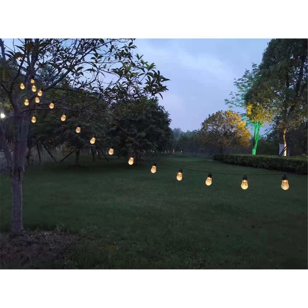 Die Solar-LED-Lichterkette mit Glühbirnen von Cover-Discount hängt in der Abenddämmerung zwischen den Bäumen in einem grasbewachsenen Hof, erfüllt den Garten mit buntem Licht und wirft einen warmen, einladenden Schein auf die Bäume.