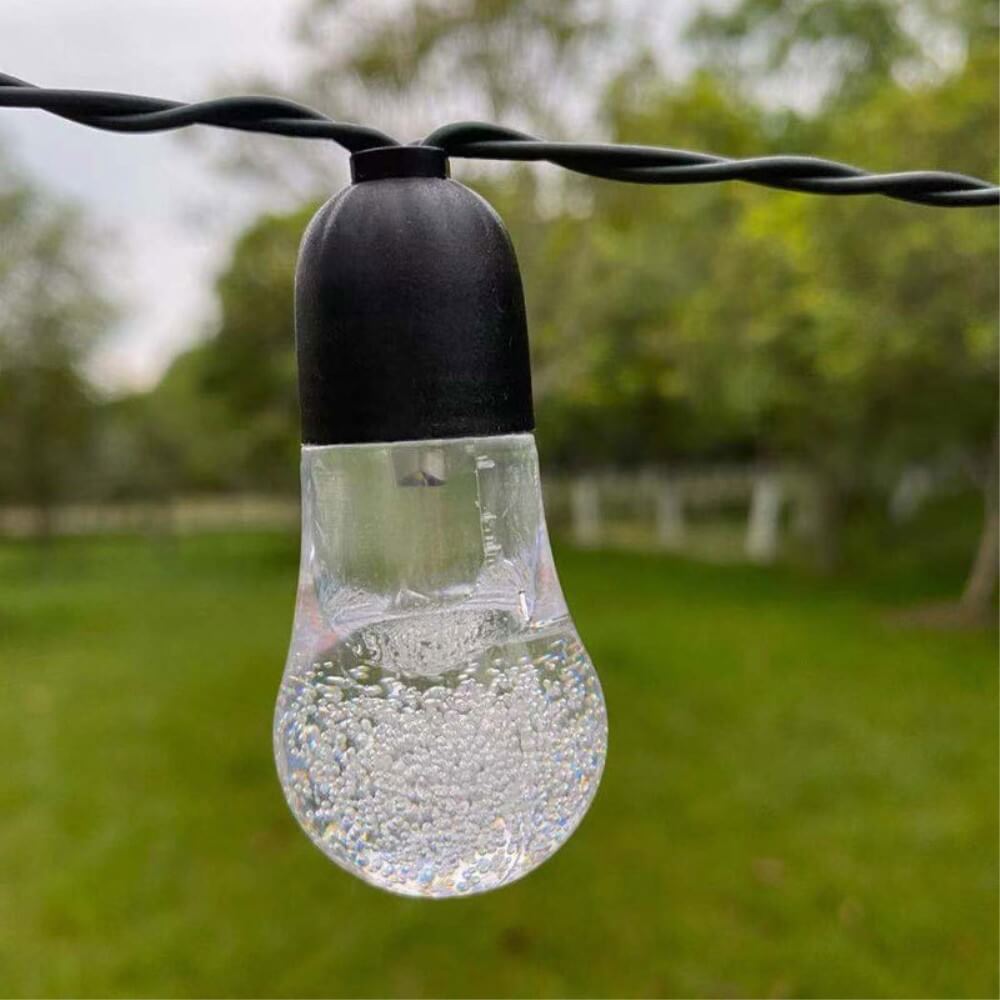 Nahaufnahme einer klaren Solar-LED-Lichterkette Glühbirnen von Cover-Discount mit Blasendesign, die in einem grünen Garten hängt – ideal für Gartenbeleuchtung mit warmem LED-Licht.
