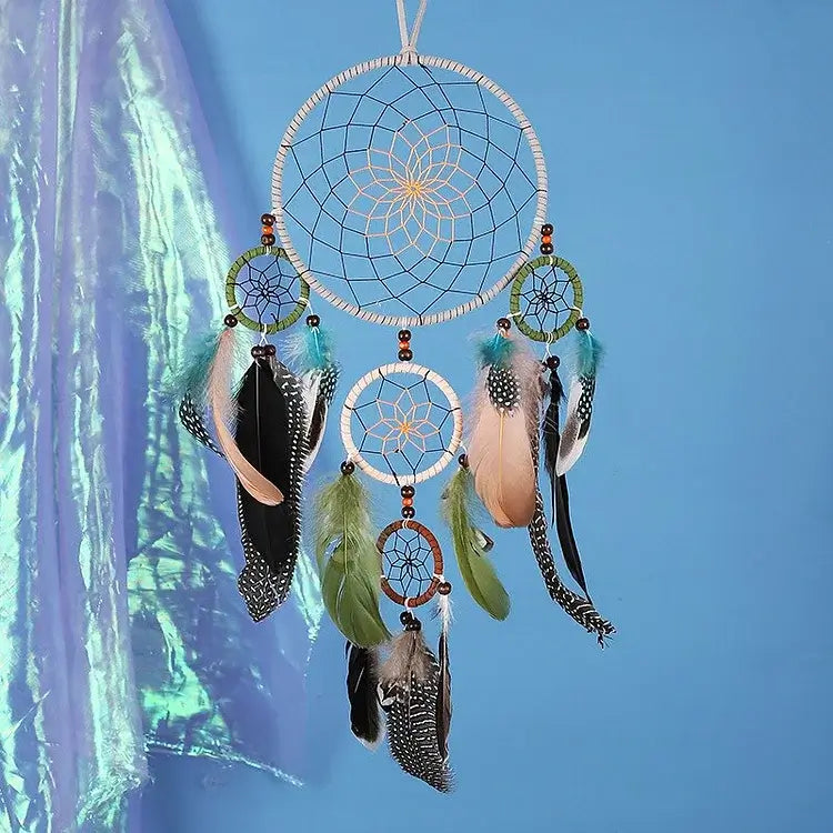 Der stilvolle Traumfänger mit 5 Ringen in Naturtönen von Cover-Discount, verziert mit bunten Federn und Perlen, ist ein einzigartiges Dekostück vor blauem Hintergrund und irisierendem Stoff.