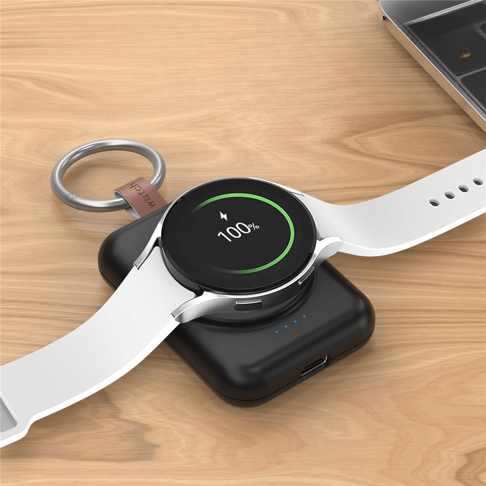 Eine weisse Smartwatch, die zu 100% aufgeladen ist, liegt auf einem schwarzen kabellosen Ladepad neben einer Cover-Discount.ch Mini Powerbank Samsung Galaxy Watch 1800mAh auf einer Holzoberfläche.