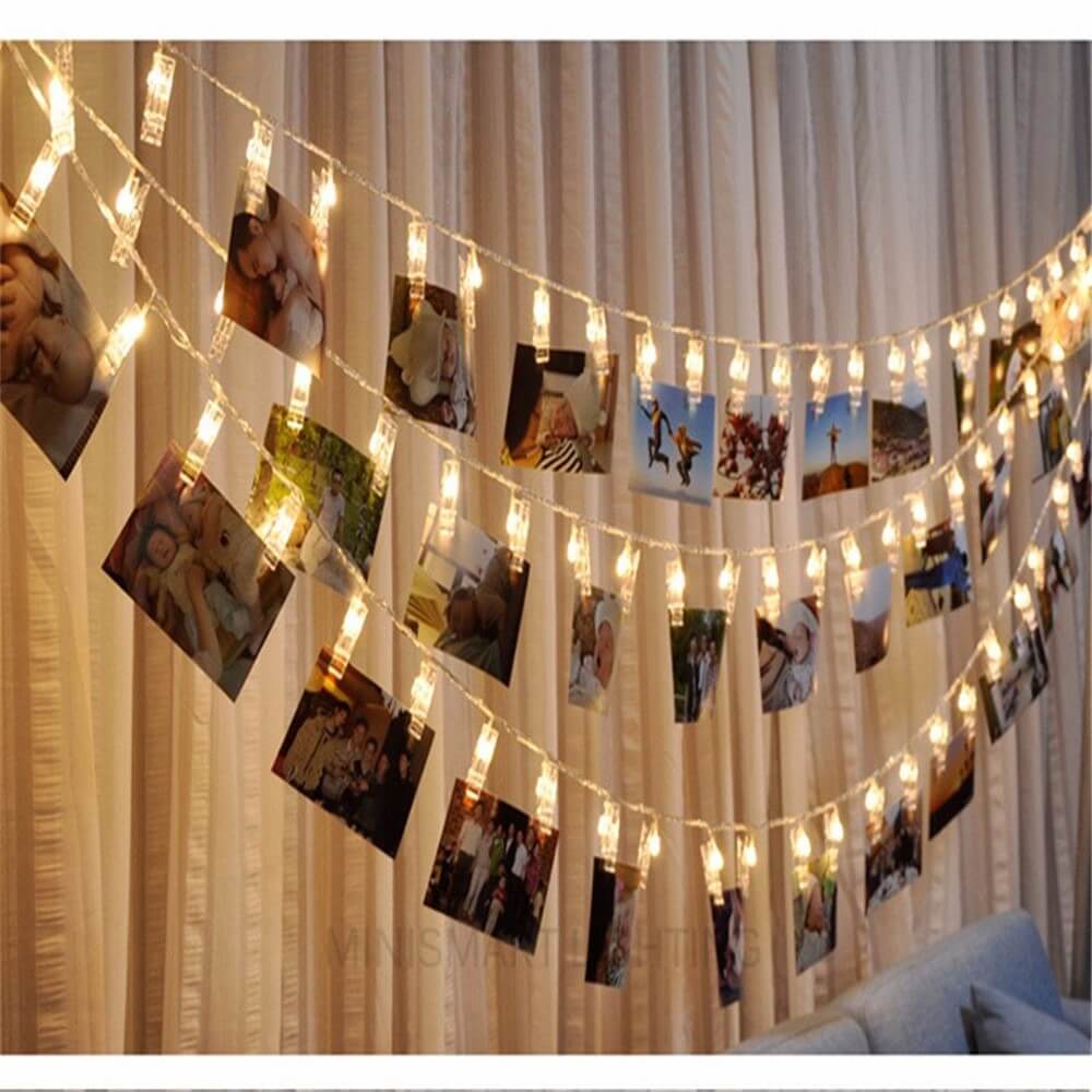 Die Lichterkette LED Fotoklammer, batteriebetrieben, von Cover-Discount zeigt kleine Fotos über einem Sofa auf beigen Vorhängen. Diese LED-Lichterkette schafft eine warme, gemütliche Atmosphäre und personalisiert Ihren Wohnraum mit beleuchteten Erinnerungen.