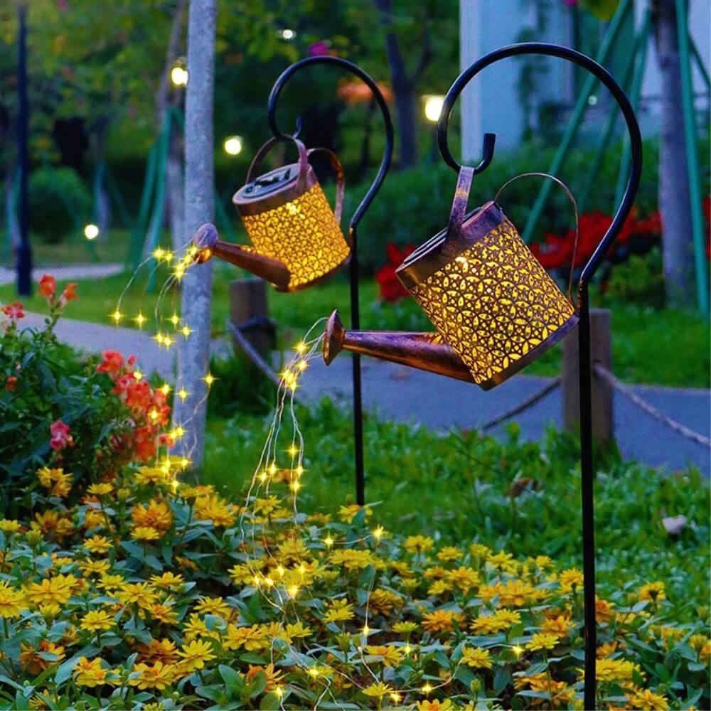 Zwei Cover-Discount Solar LED Gartenstecker Giesskanne scheinen warmweißes Licht über gelbe Blumen zu gießen und erzeugen so einen dekorativen goldenen Schimmer im Garten.