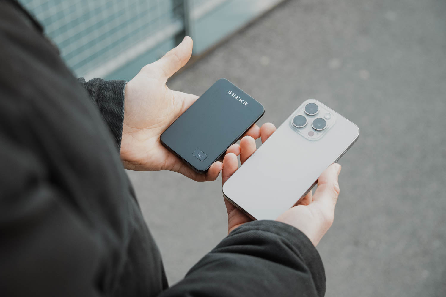 Eine Person hält das SEEKR SWISS SEEKR Power Mag - Schwarz 5000 mAh mit ultraschlankem Design in einer Hand und ein silbernes Smartphone in der anderen, während sie im Freien ist.